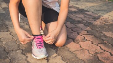 Photo of a woman kneeling down to tie her shoe. You cannot see her head/face, but the shoe has pink shoelaces. This is to illustrate an article titled 4 Summer Fitness Tips to Help You Get Outside