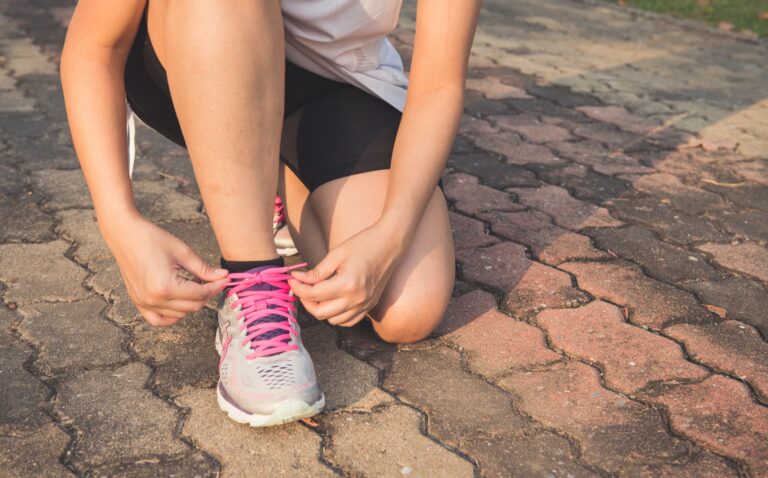Photo of a woman kneeling down to tie her shoe. You cannot see her head/face, but the shoe has pink shoelaces. This is to illustrate an article titled 4 Summer Fitness Tips to Help You Get Outside